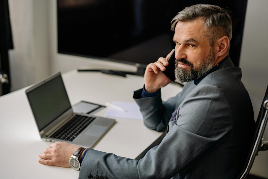 Confident mature businessman talking on phone at desk with laptop.